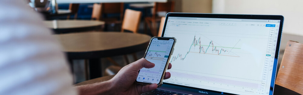 Eine Person sitzt an einem Tisch mit einem Laptop und hält ein Smartphone in der Hand. Der Laptop zeigt ein Diagramm mit Kursmustern und Trendlinien.