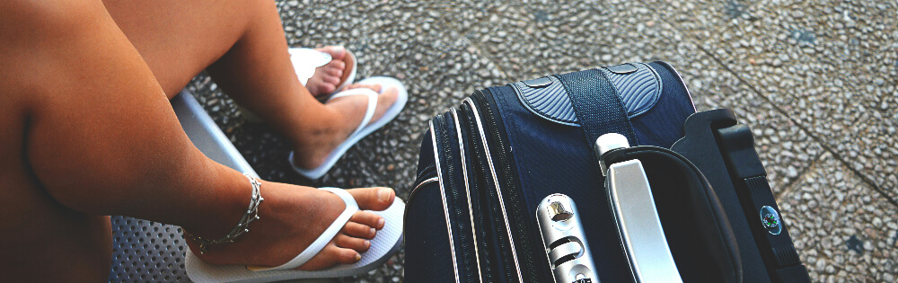 Person sitzt auf einer Bank mit einem schwarzen Reisekoffer daneben und trägt weiße Flip-Flops sowie ein Fußkettchen.