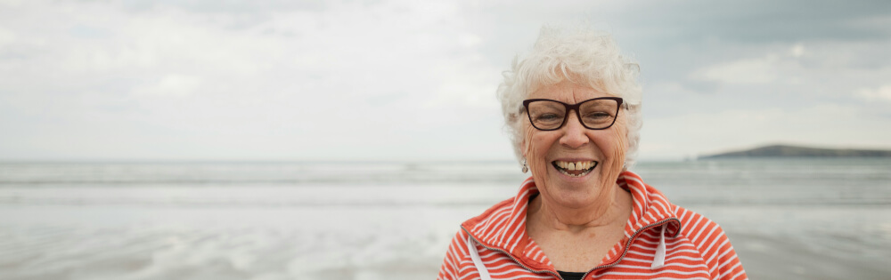 Eine ältere Frau mit weißem Haar und Brille lächelt fröhlich am Strand. Sie trägt ein gestreiftes Oberteil und im Hintergrund ist das Meer mit einem wolkigen Himmel zu sehen.